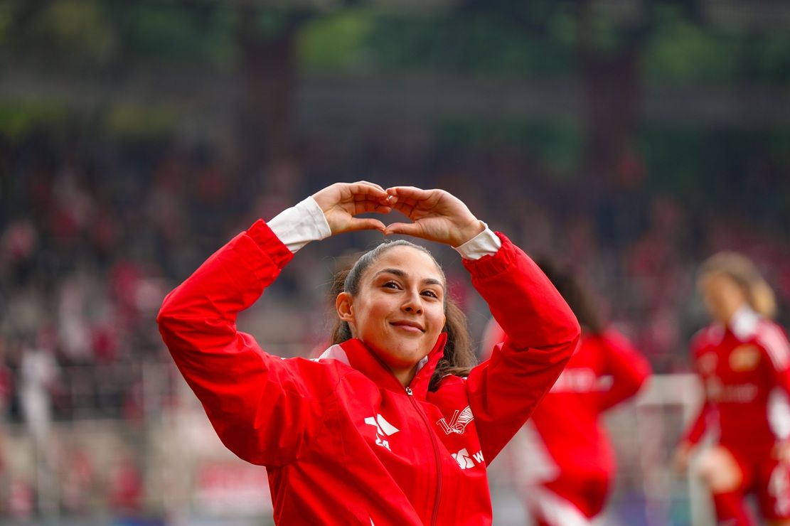 Eine sportliche Frau in roter Jacke macht ein Herz mit ihren Händen, im Hintergrund ein unscharfes Stadion.