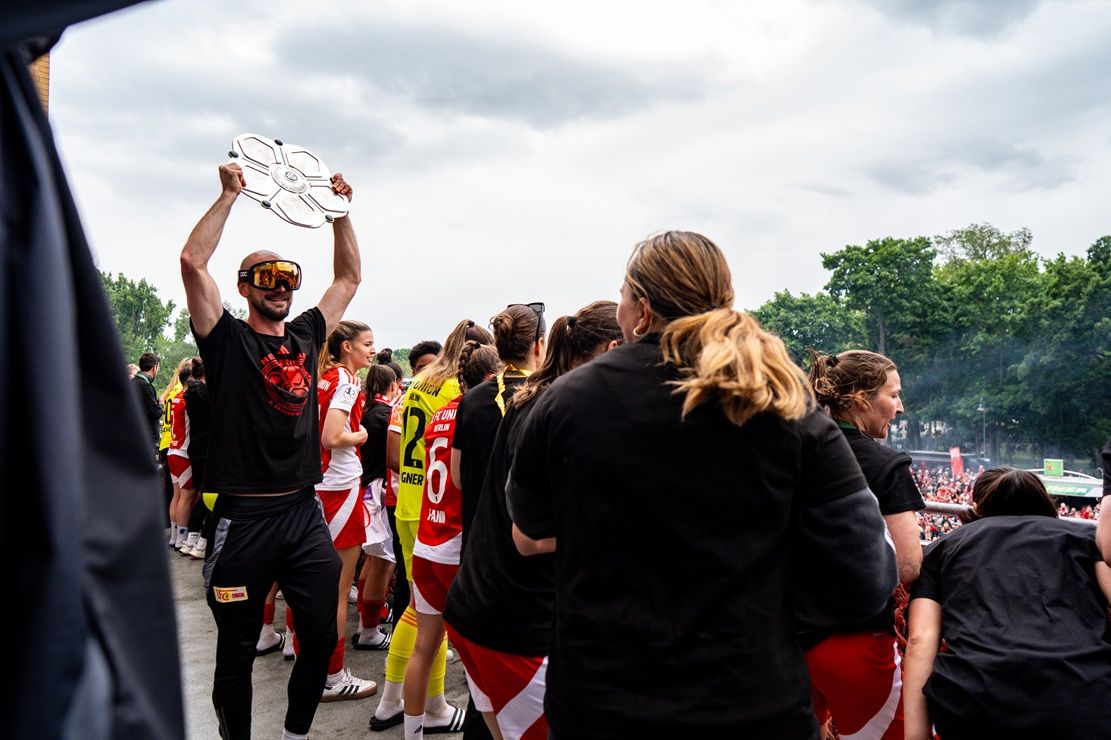 Ein Team feiert mit Freude, während ein Spieler den Pokal in die Höhe hält; umgeben von jubelnden Kolleginnen.