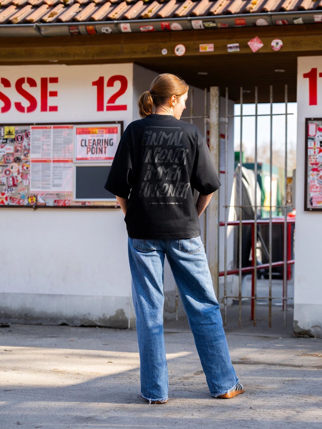 Eine Person steht mit dem Rücken zur Kamera vor einem Eingang, trägt ein schwarzes Shirt und weite Jeans.