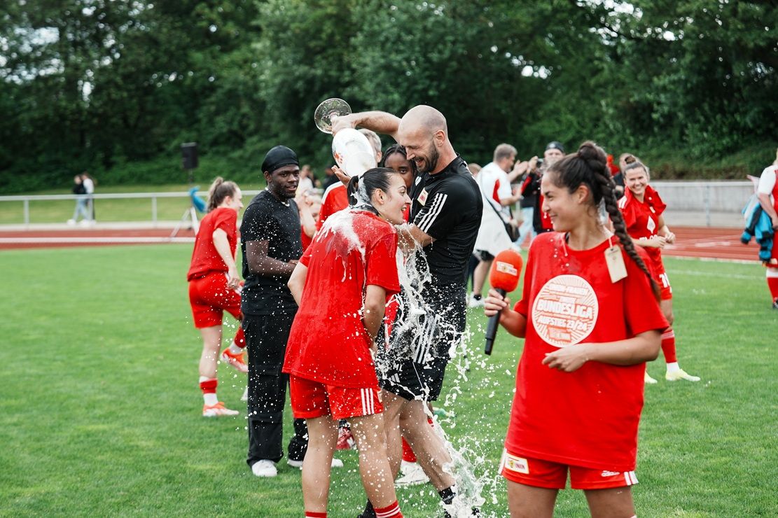 Eine Gruppe von Fußballspielerinnen feiert mit Sprudelwasser auf einem Rasenplatz, während einige lächeln und jubeln.