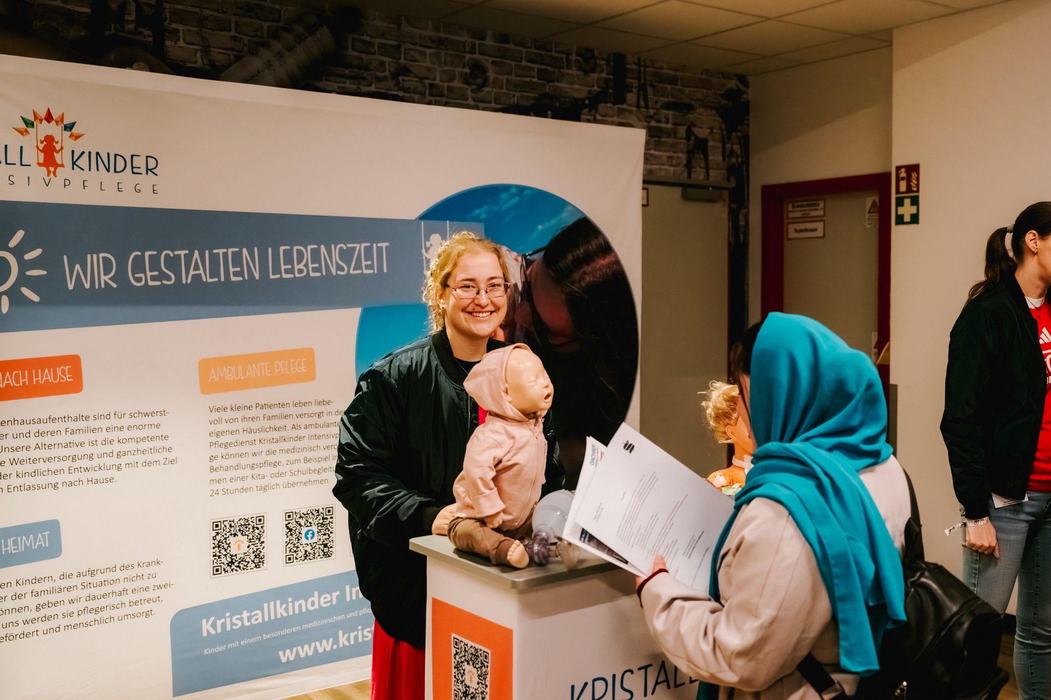 Eine Person spricht an einem Informationsstand mit einer anderen, während eine Puppe auf dem Tisch steht.