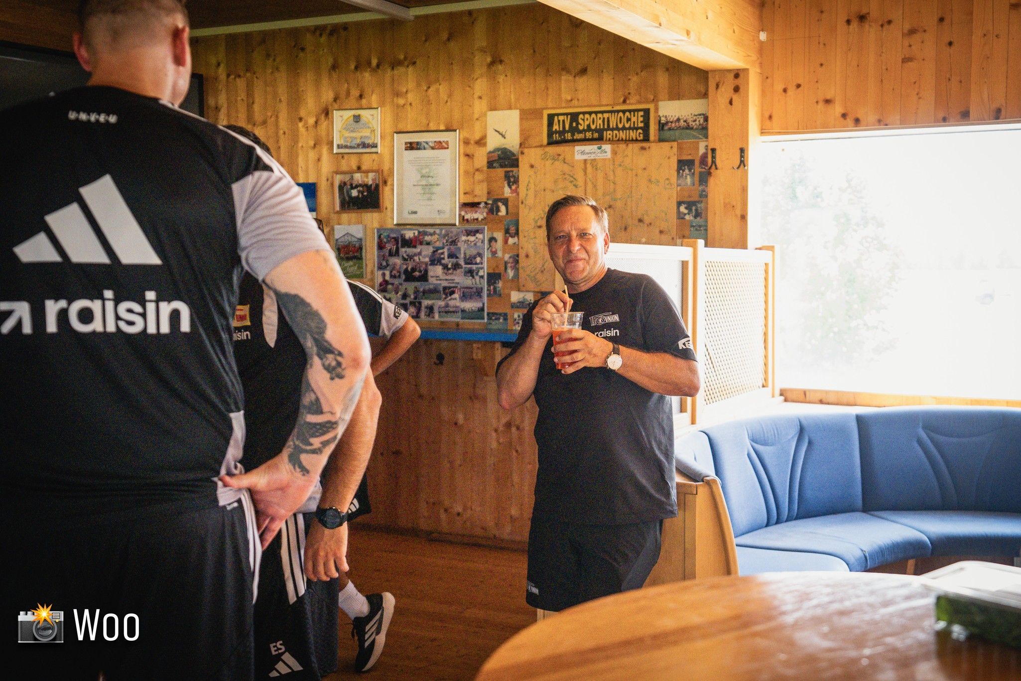 Ein Mann in schwarzem Shirt hält ein Getränk und steht in einem Holzraum, umgeben von weiteren Personen.
