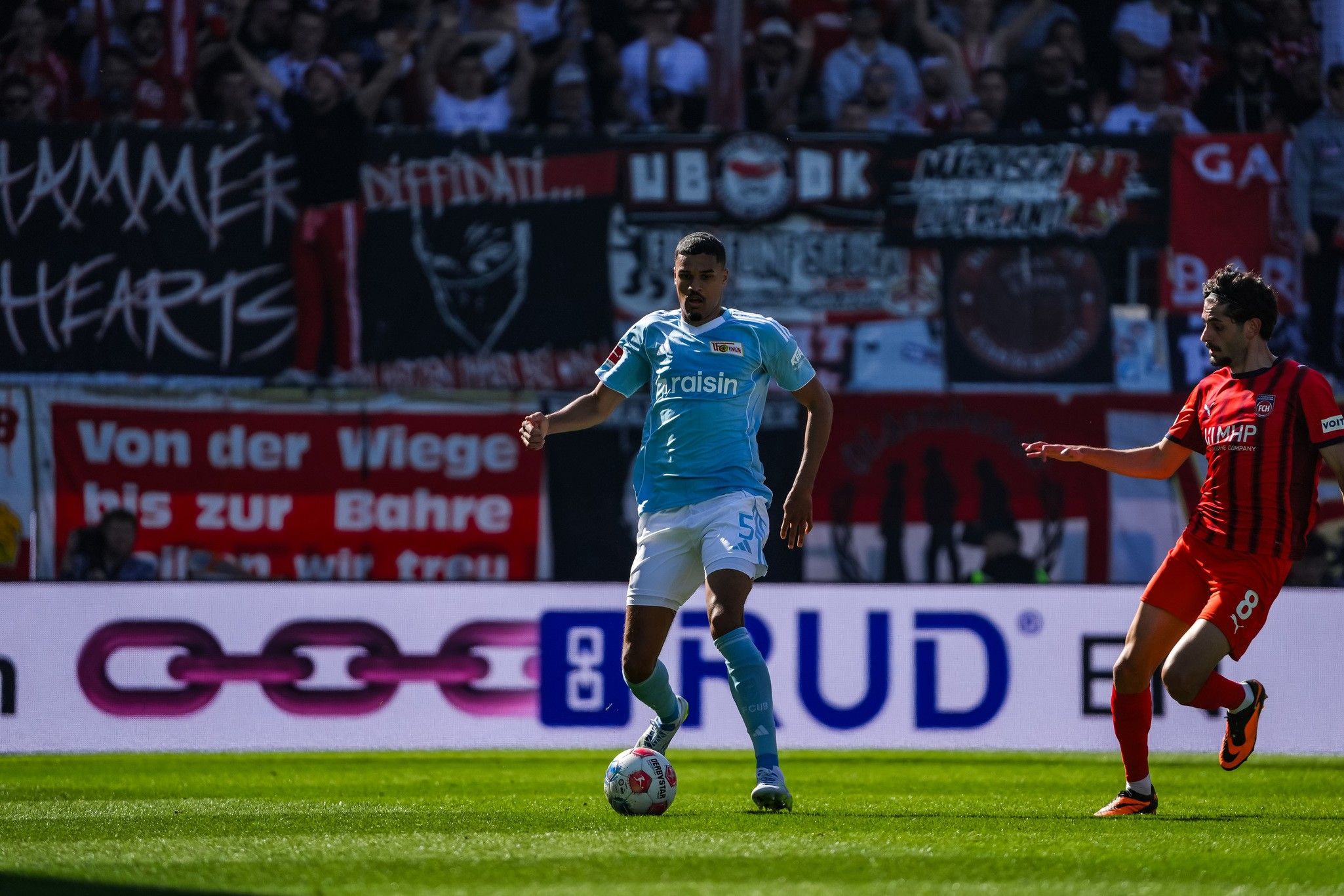 Ein Fußballspieler in hellblauem Trikot steht auf dem Spielfeld, während ein Gegner in dunklem Trikot in seiner Nähe läuft. Fans mit Fahnen im Hintergrund.