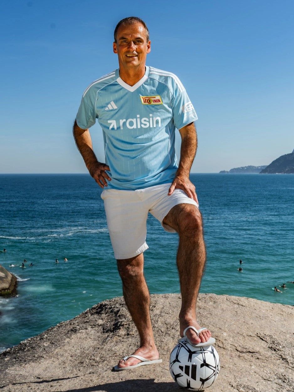 Mann im hellblauen Fußballtrikot und Shorts steht am Strand auf einer Felsplatte neben einem Fußball, mit Meer im Hintergrund.