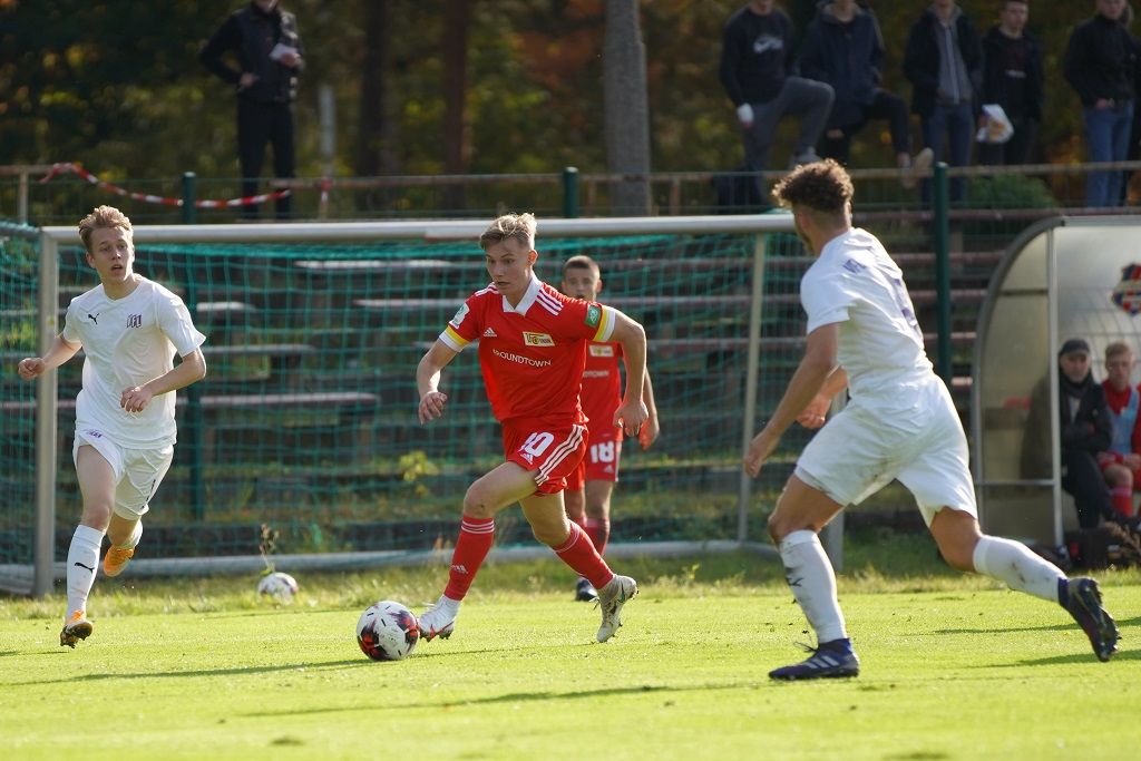 Ein junger Fußballspieler in rotem Trikot dribbelt den Ball, während zwei Gegner in weißen Trikots versuchen, ihn zu stoppen.