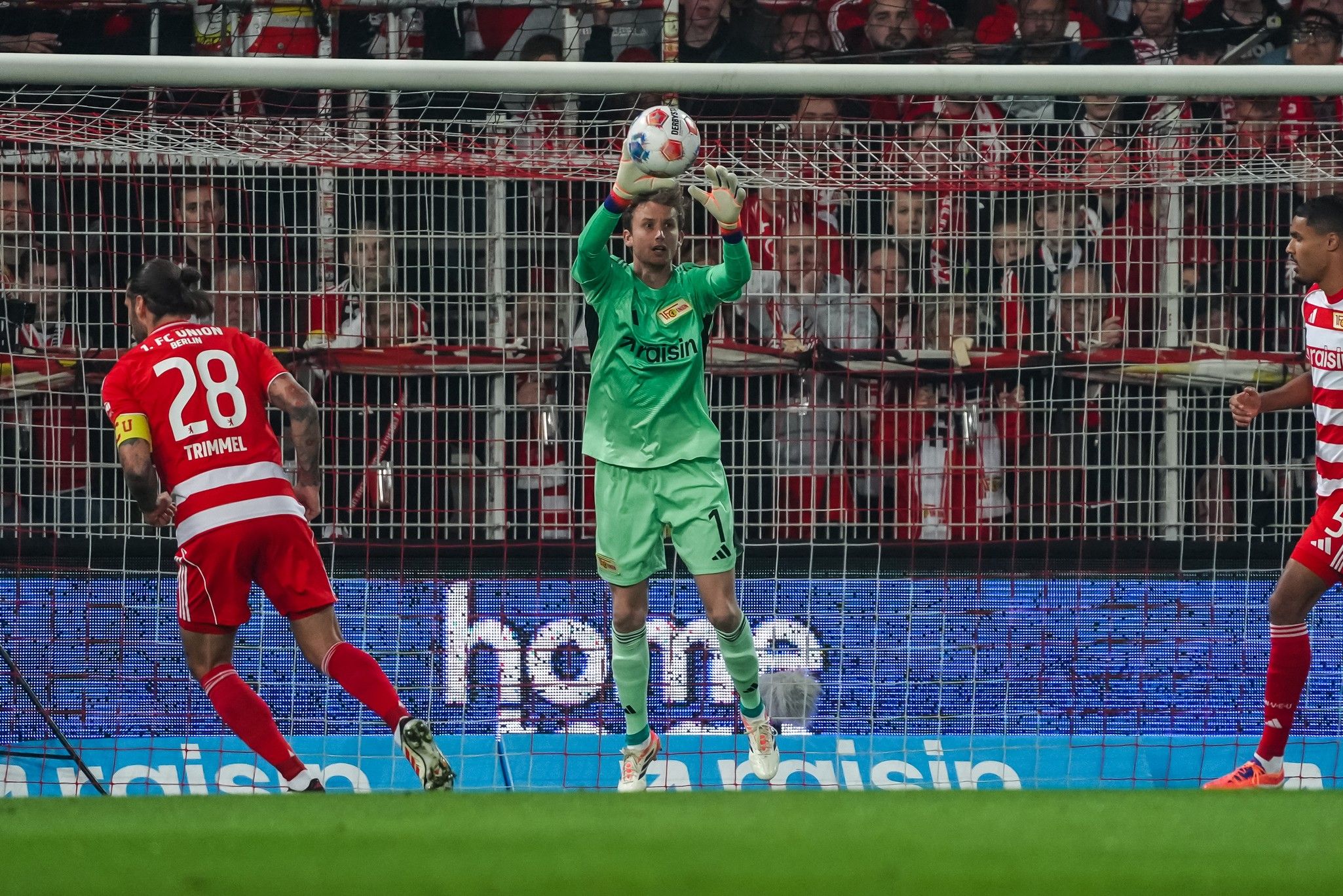 Torwart hält den Ball in einem Fußballspiel, während ein Spieler in Rot versucht, ihn zu überwinden. Fans im Hintergrund sind zu sehen.