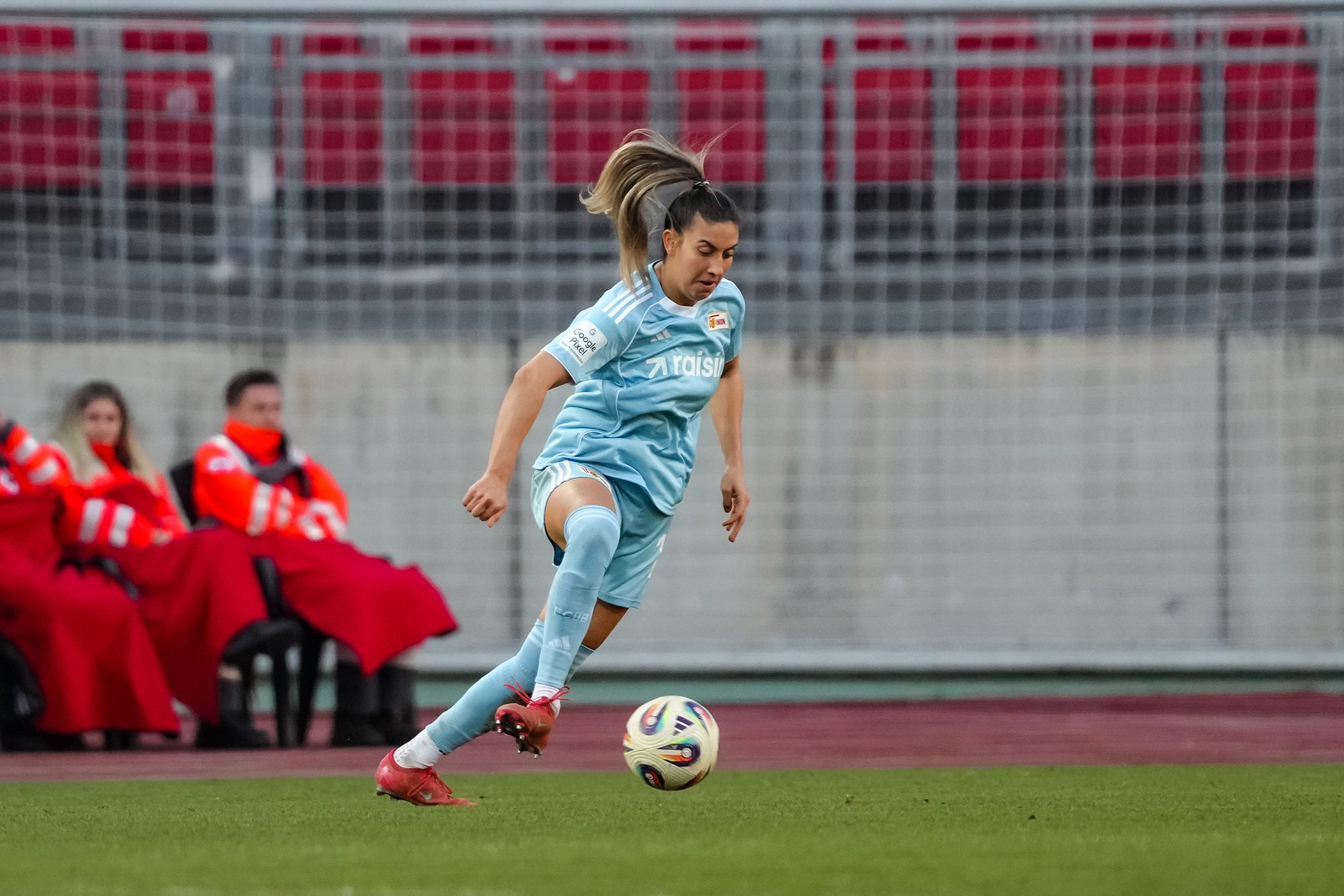 Eine Fußballspielerin in blauer Ausrüstung tritt den Ball während eines Spiels. Im Hintergrund sitzen Zuschauer in roten Decken.