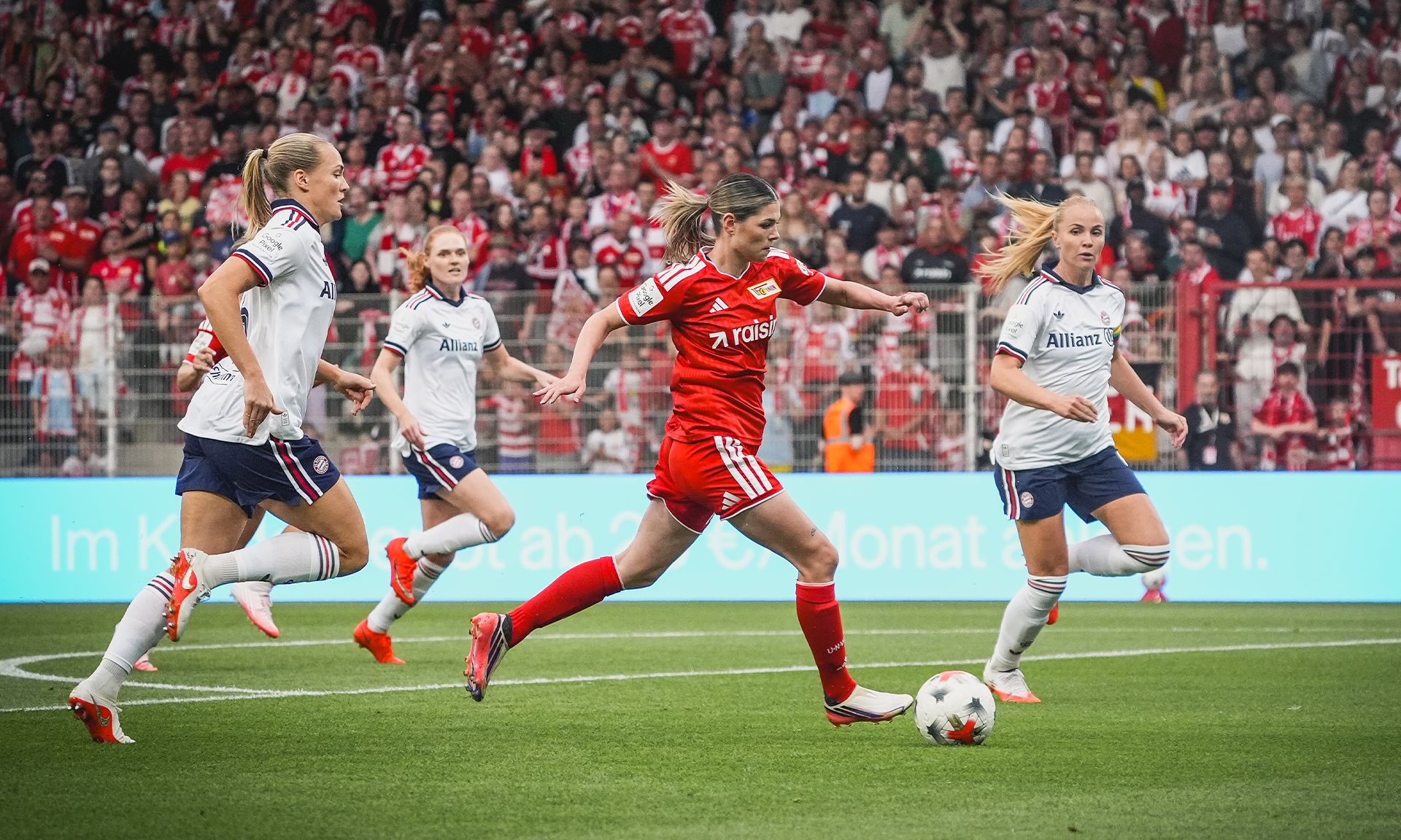 Eine Spielerin in rotem Trikot sprintet mit Ball, umgeben von zwei Gegenspielerinnen in weißen Trikots, im Stadion voller Zuschauer.