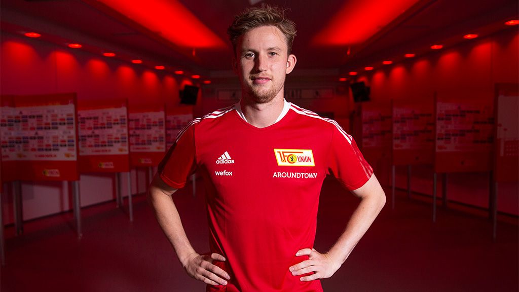 A young man in a red jersey with white details stands in a brightly lit room with red walls.