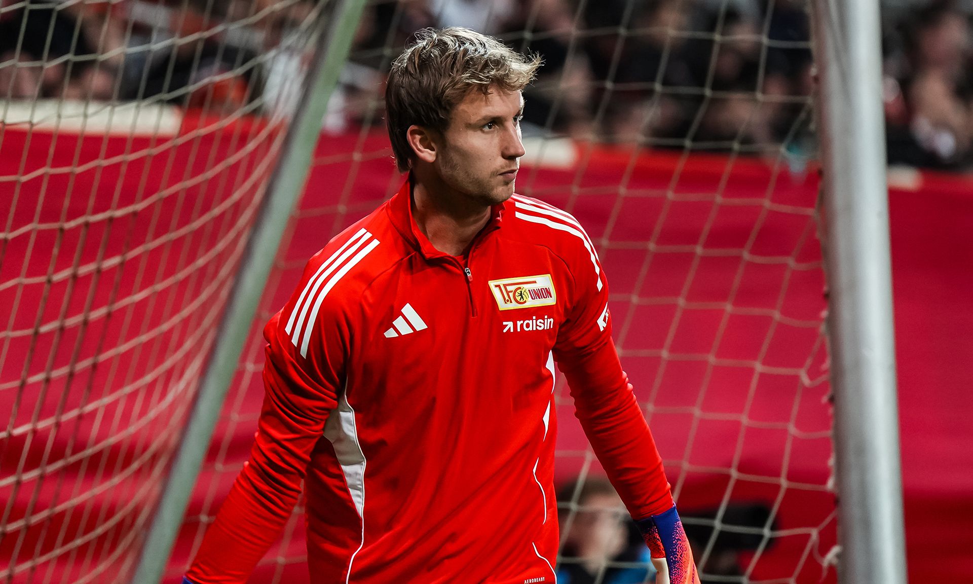 Torhüter in rotem Trikot steht im Tor, Blick konzentriert auf das Spiel, im Hintergrund verschwommene Zuschauer und Stadionbeleuchtung.