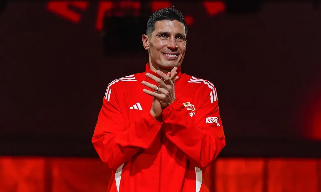 Ein Mann in rot-orangefarbener Sportjacke steht lächelnd und applaudiert, umgeben von einem dunklen Hintergrund mit Lichtakzenten.
