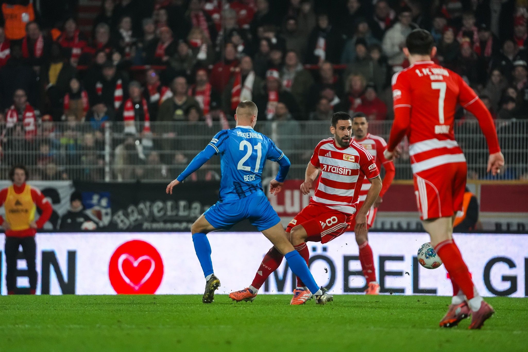 Zwei Fußballspieler kämpfen um den Ball in einem Stadion, während Zuschauer im Hintergrund zu sehen sind.