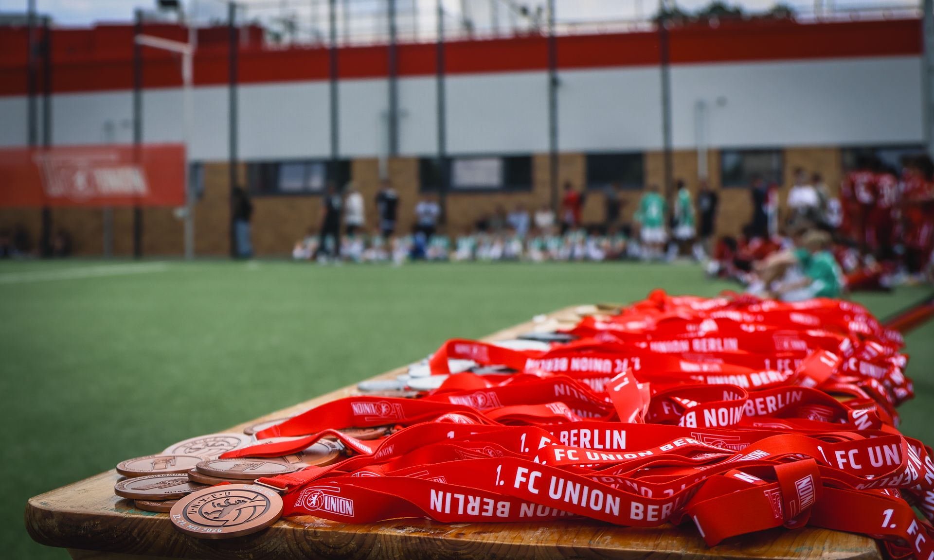 Rote Medaillen mit dem Aufdruck "1. FC UNION BERLIN" liegen auf einem Tisch, im Hintergrund sind Spieler und Zuschauer zu sehen.