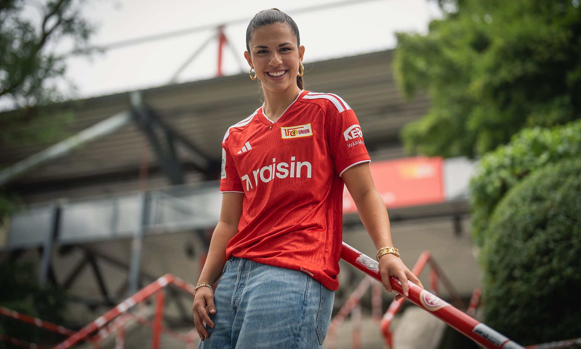 Frau in rotem Fußballtrikot steht lächelnd vor einem Stadion und posiert für das Foto.