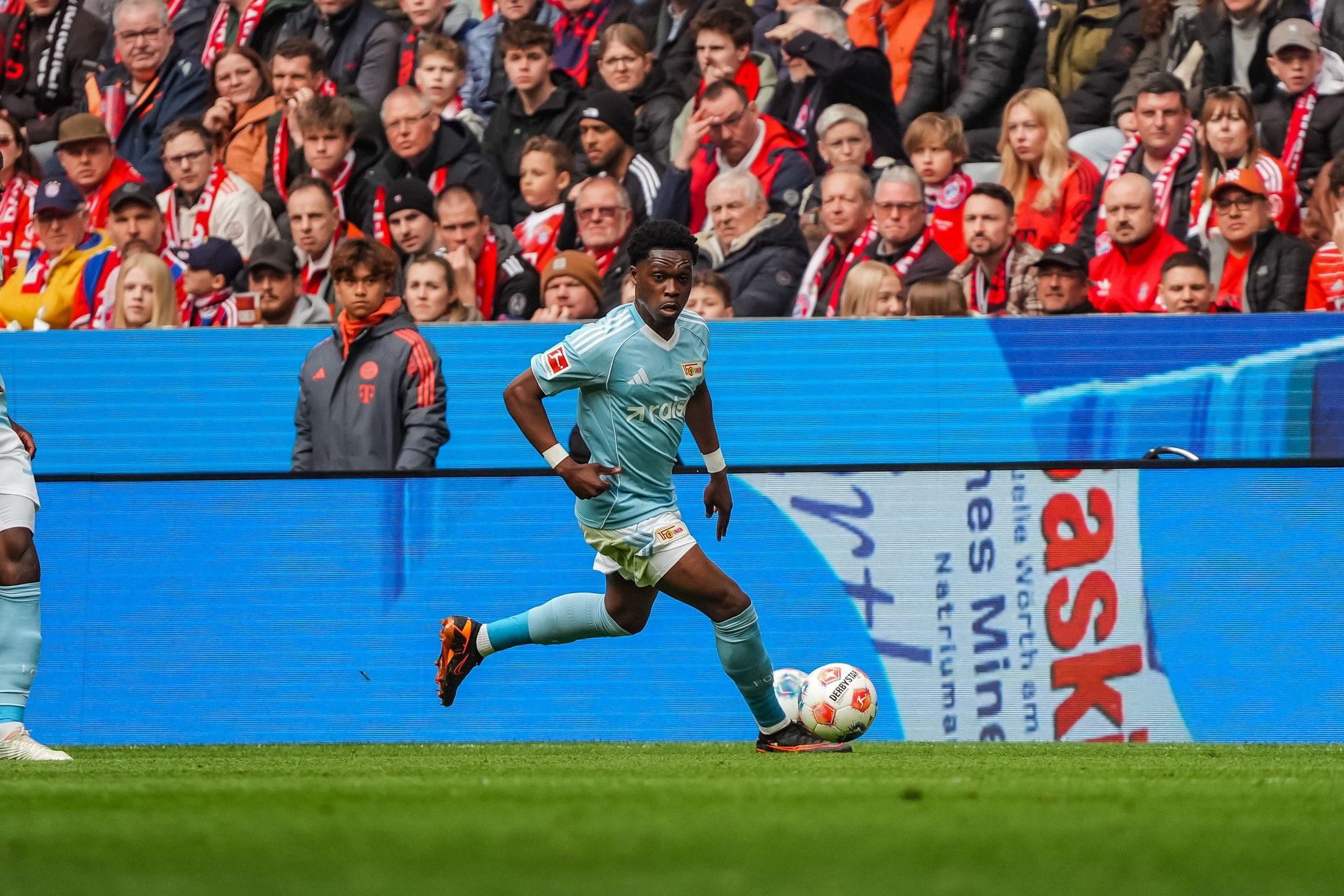 Ein Fußballspieler in hellblauem Trikot dribbelt den Ball auf einem Spielfeld, während Zuschauer im Hintergrund zuschauen.