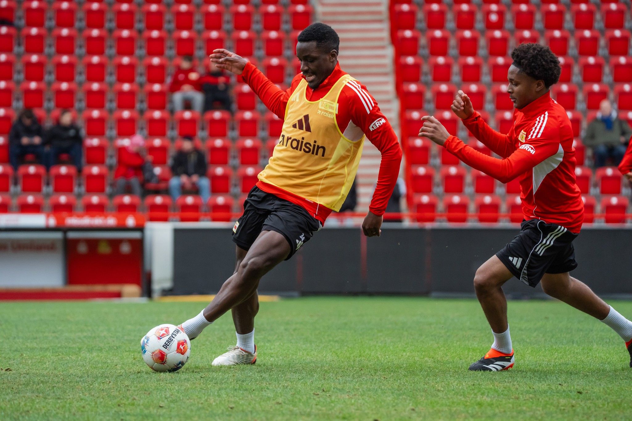 Zwei Fußballspieler in Trainingskleidung üben auf einem Stadionrasen, im Hintergrund sind leere Sitzplätze sichtbar.