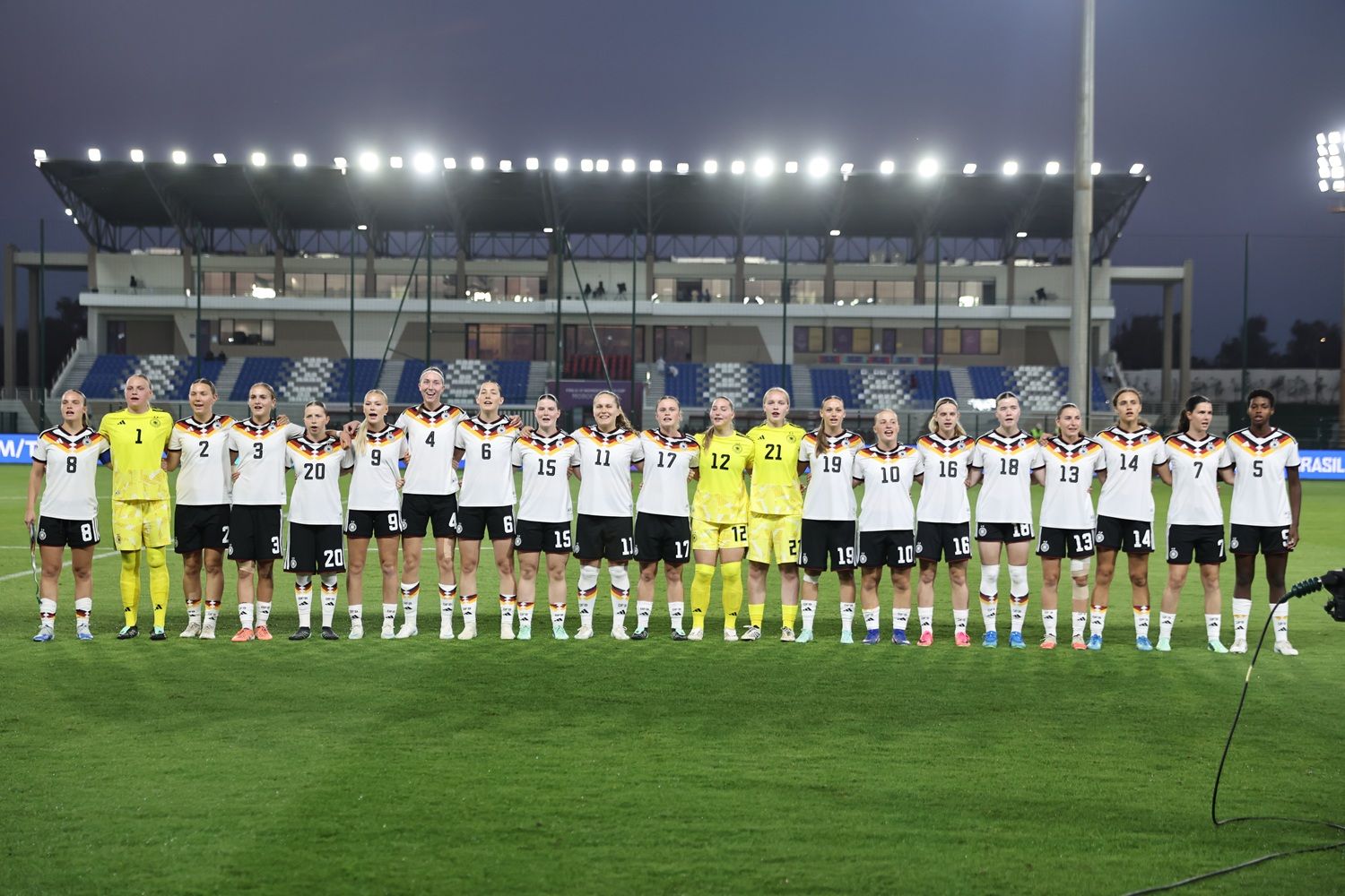 Die deutsche Frauenfußballmannschaft steht in zwei Reihen auf dem Platz, in einheitlichen Trikots, bei Dämmerung.