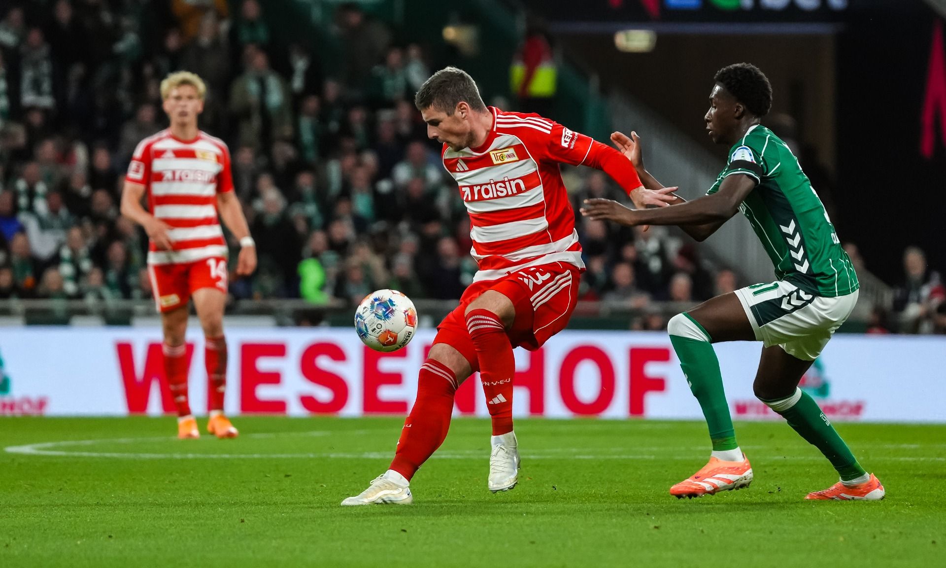 Spielszene mit einem Fußballspieler in roter Vereinskleidung, der einen gegnerischen Spieler in grüner Kleidung dribbelt.