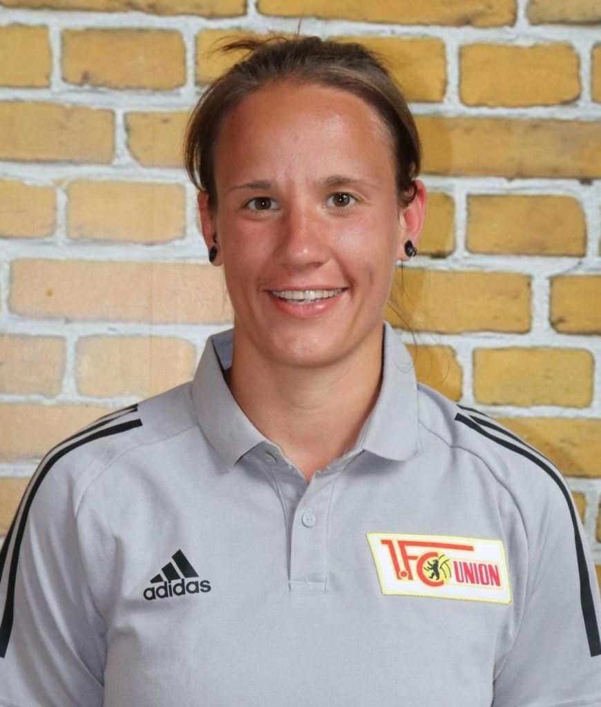 Porträt einer Person in einem grauen Sporthemd mit dem Logo von 1. FC Union Berlin und einer Backsteinmauer im Hintergrund.
