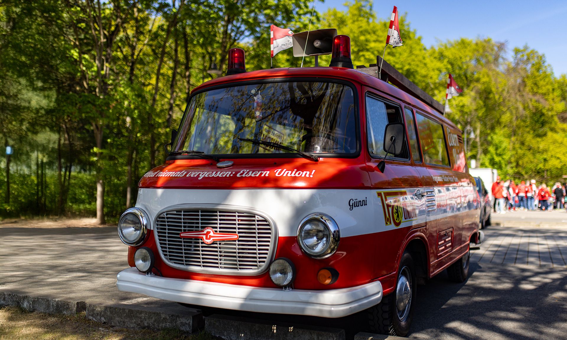 Oldtimer-Lkw in rot-weiß mit Sirene und Flaggen, abgestellte auf einem Waldweg, umgeben von Bäumen und Menschen im Hintergrund.