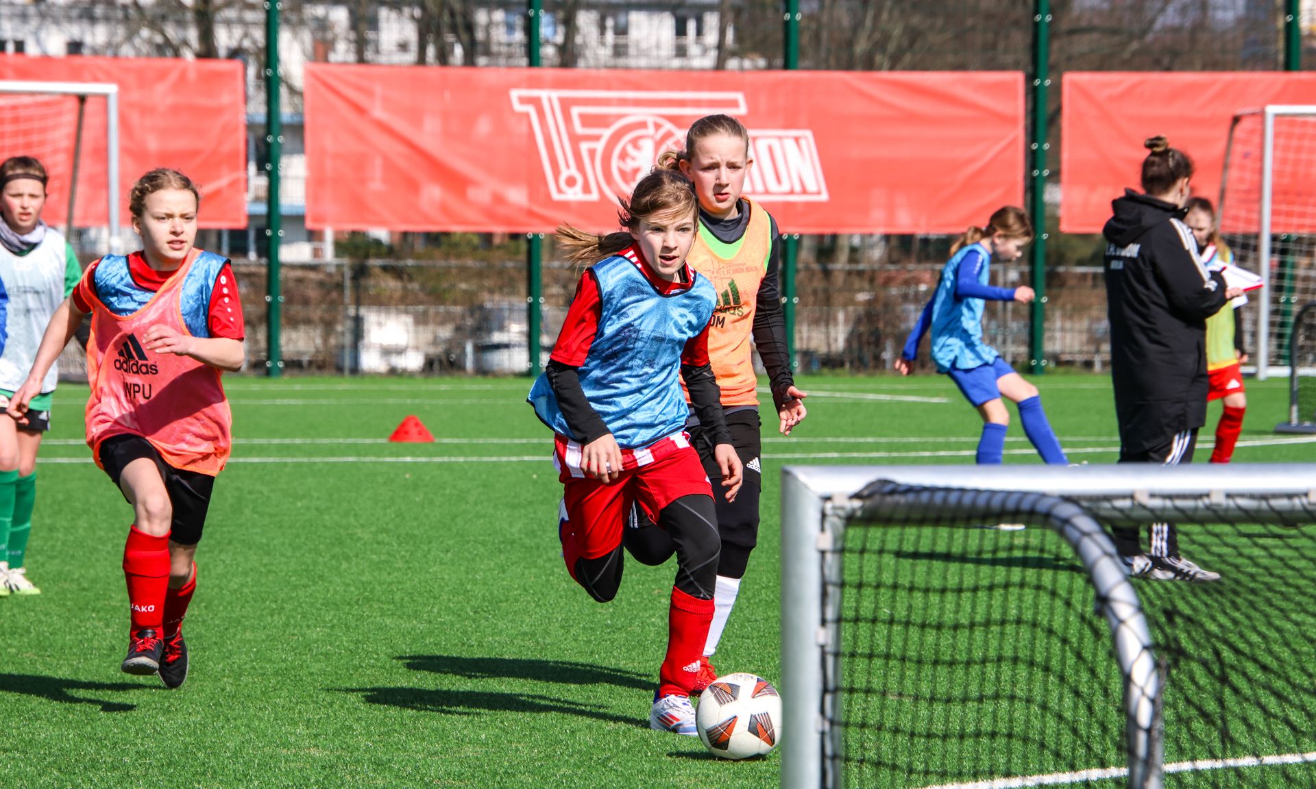 Mädchen in Fußballtrikots trainieren auf einem Platz, während sie einen Ball Richtung Tor dribbeln.
