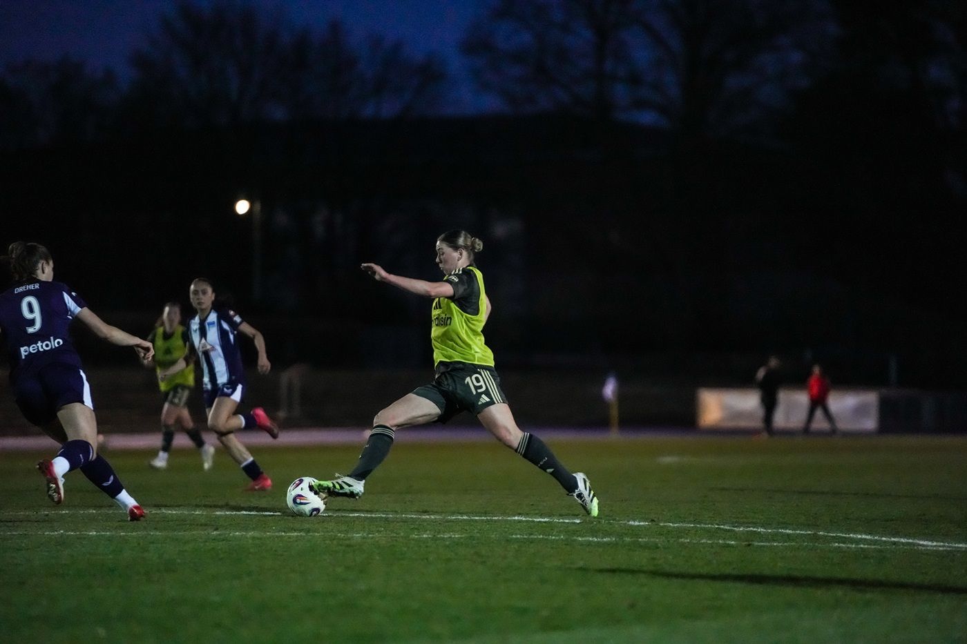 Spielerin in gelber Trikots macht einen Schuss auf dem Fußballplatz bei Dämmerung, umgeben von anderen Spielerinnen.