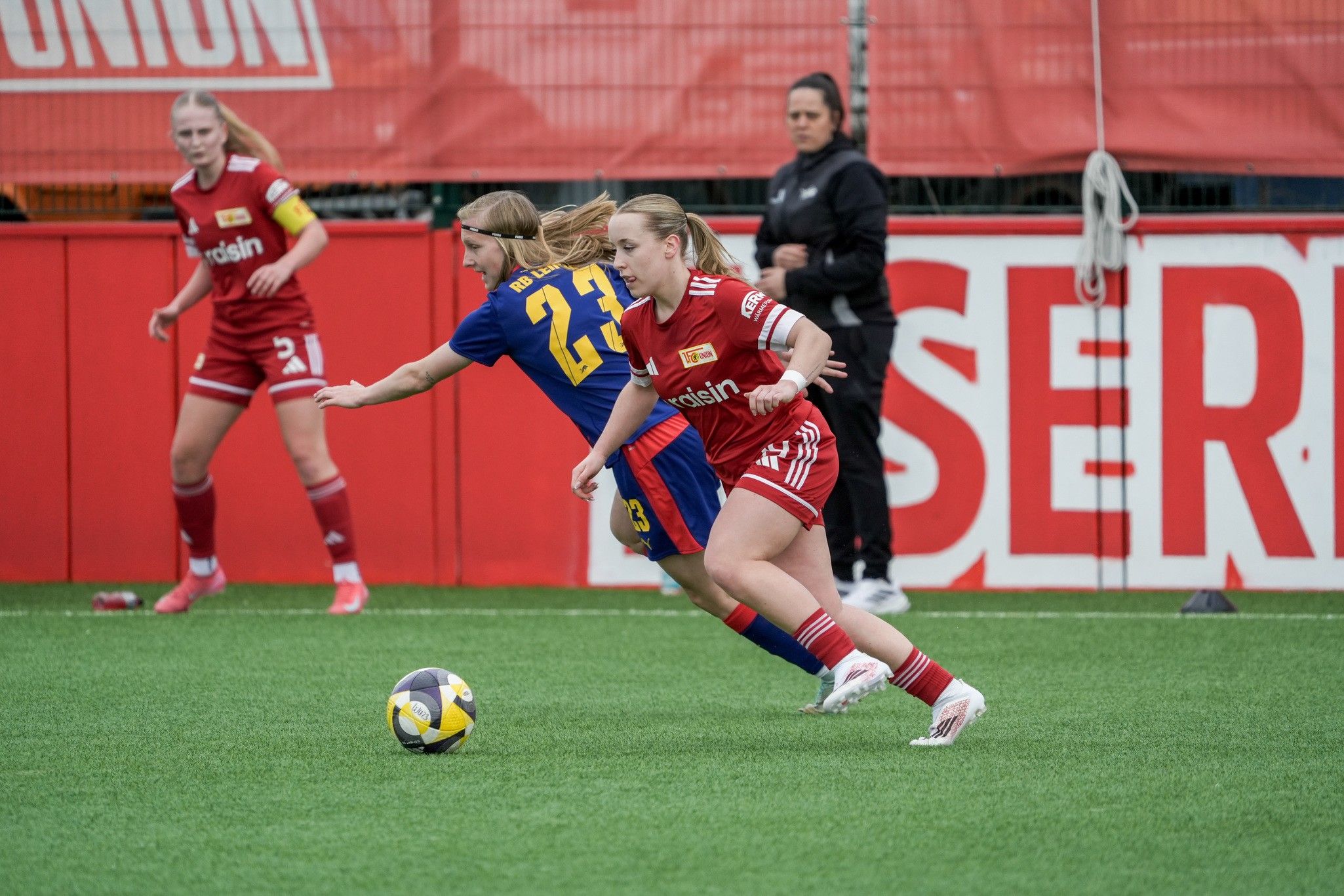 Zwei Fußballspielerinnen im Wettlauf um den Ball auf einem grünen Spielfeld, im Hintergrund Zuschauer und Trainer.