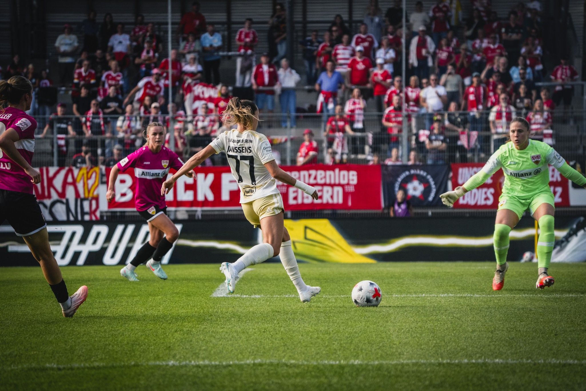 Eine Spielerin schießt im Fußballspiel auf das Tor, während ein Torwart sich auf den Schuss vorbereitet, im Hintergrund jubelnde Fans.