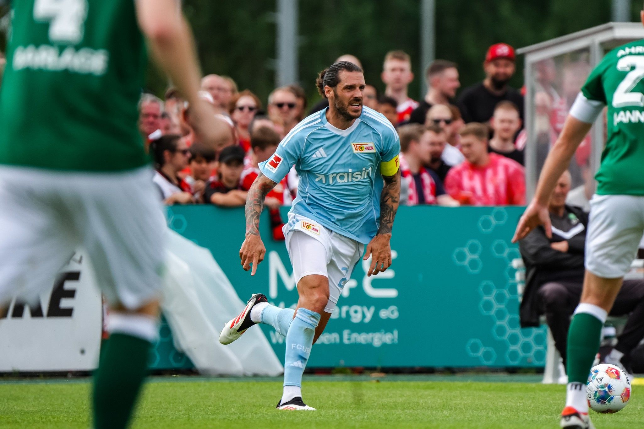 Ein Fußballspieler in hellblauem Trikot läuft mit dem Ball, während Fans im Hintergrund jubeln.