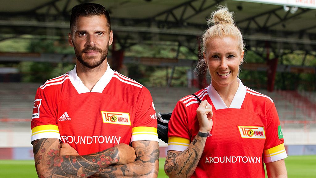 Zwei Sportler in roten Fußballtrikots stehen in einem Stadion, mit Freundlichen Gesichtsausdrücken und verschränkten Armen.