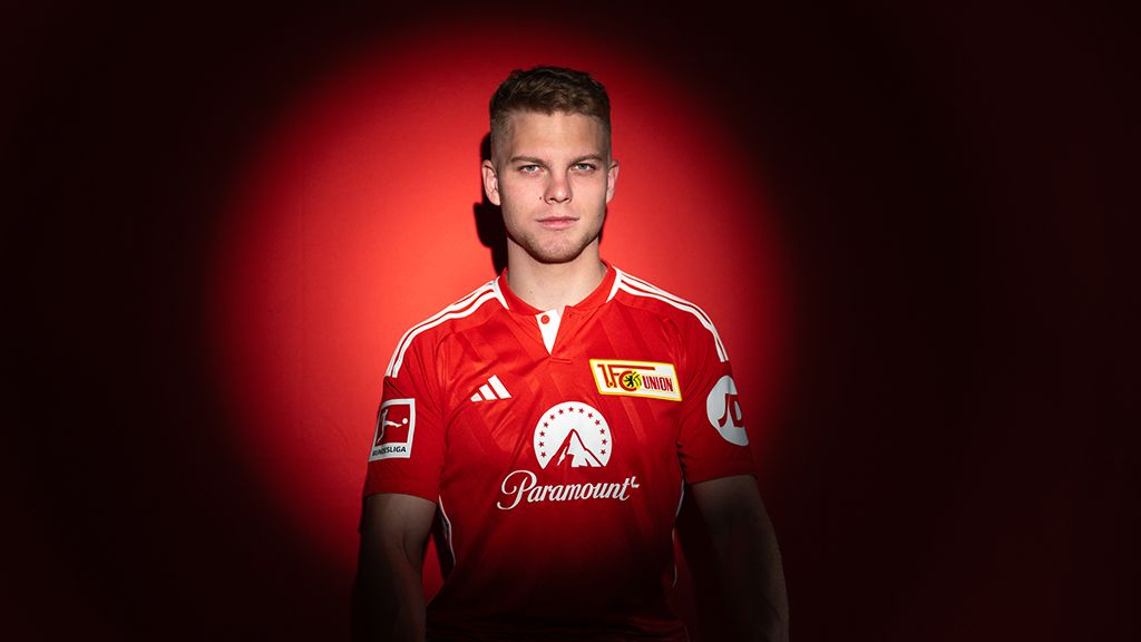 Ein junger Fußballspieler in rotem Trikot steht vor einem roten Hintergrund und schaut direkt in die Kamera.