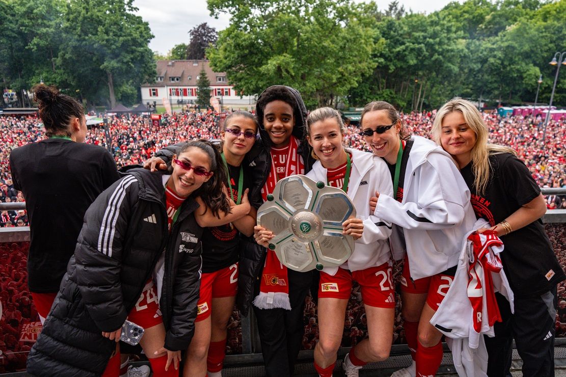 Sechs weibliche Fußballspielerinnen in roten Trikots halten einen Pokal, umgeben von einer jubelnden Menge.