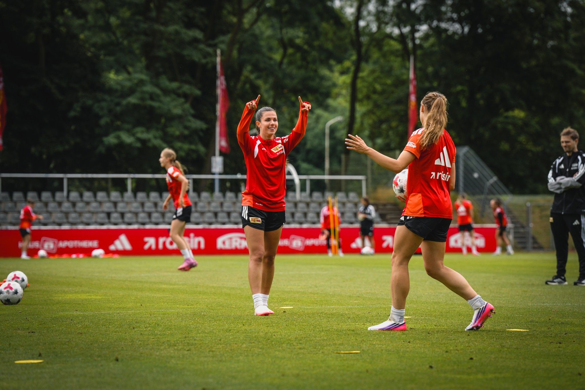 Zwei Fußballspielerinnen in roten Trainingsshirts feiern während des Trainings auf einem grünen Platz im Freien.