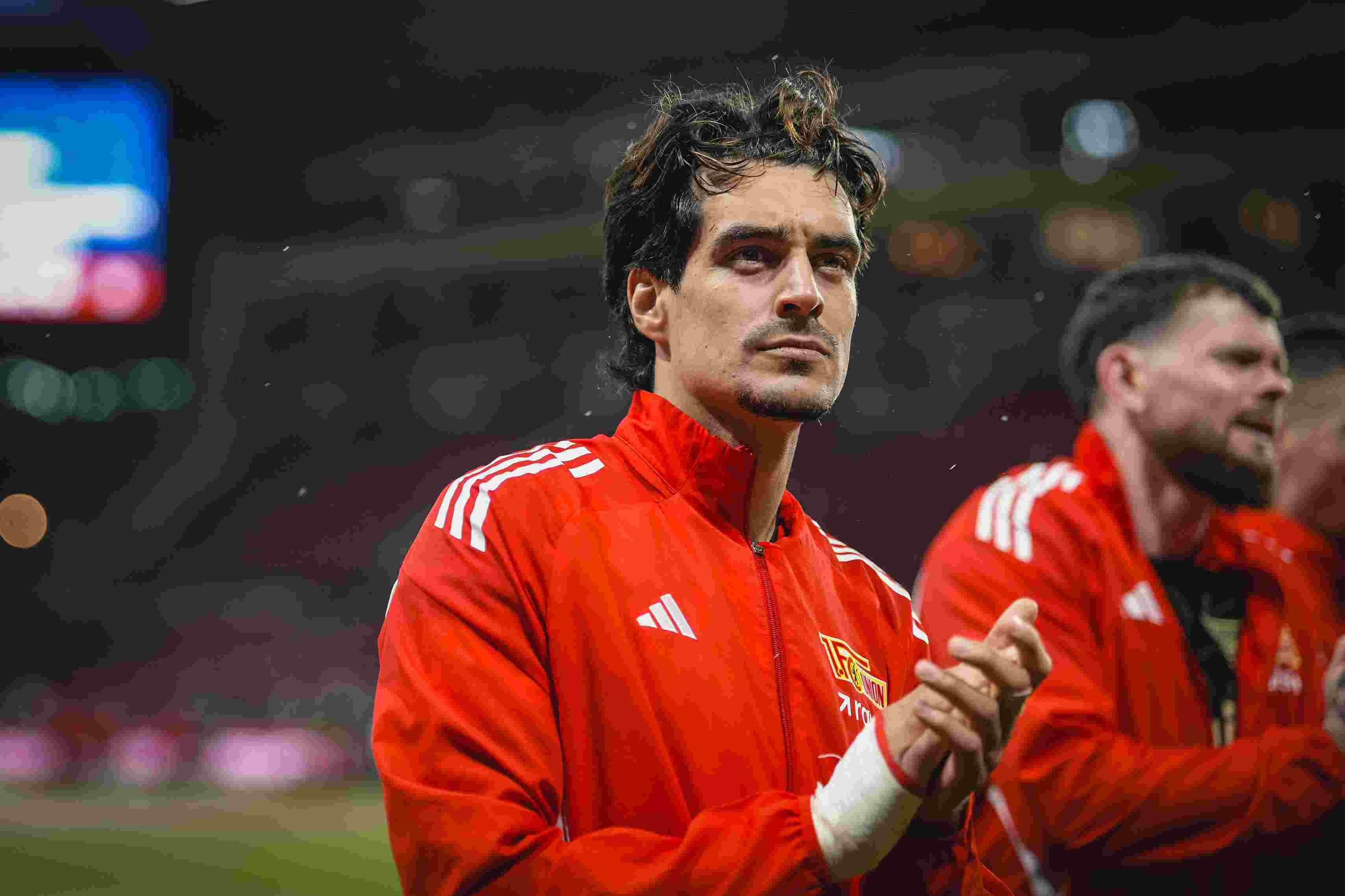 Ein Sportler in roter Trainingsjacke steht nachdenklich in einem Stadion, während andere Personen im Hintergrund applaudieren.