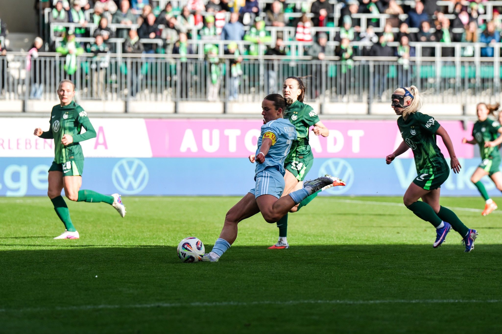 Eine Fußballspielerin in blauer Spielkleidung führt einen Schuss aus, während Spielerinnen in grüner Kleidung sich in der Nähe befinden.