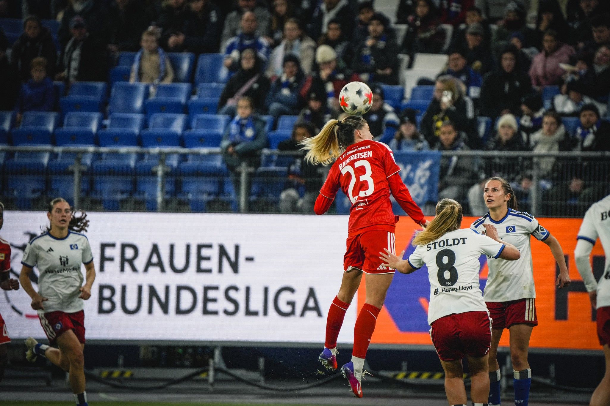 Eine Fußballspielerin springt hoch, um den Ball zu erreichen, während andere Spielerinnen in der Nähe stehen. Fans im Hintergrund.