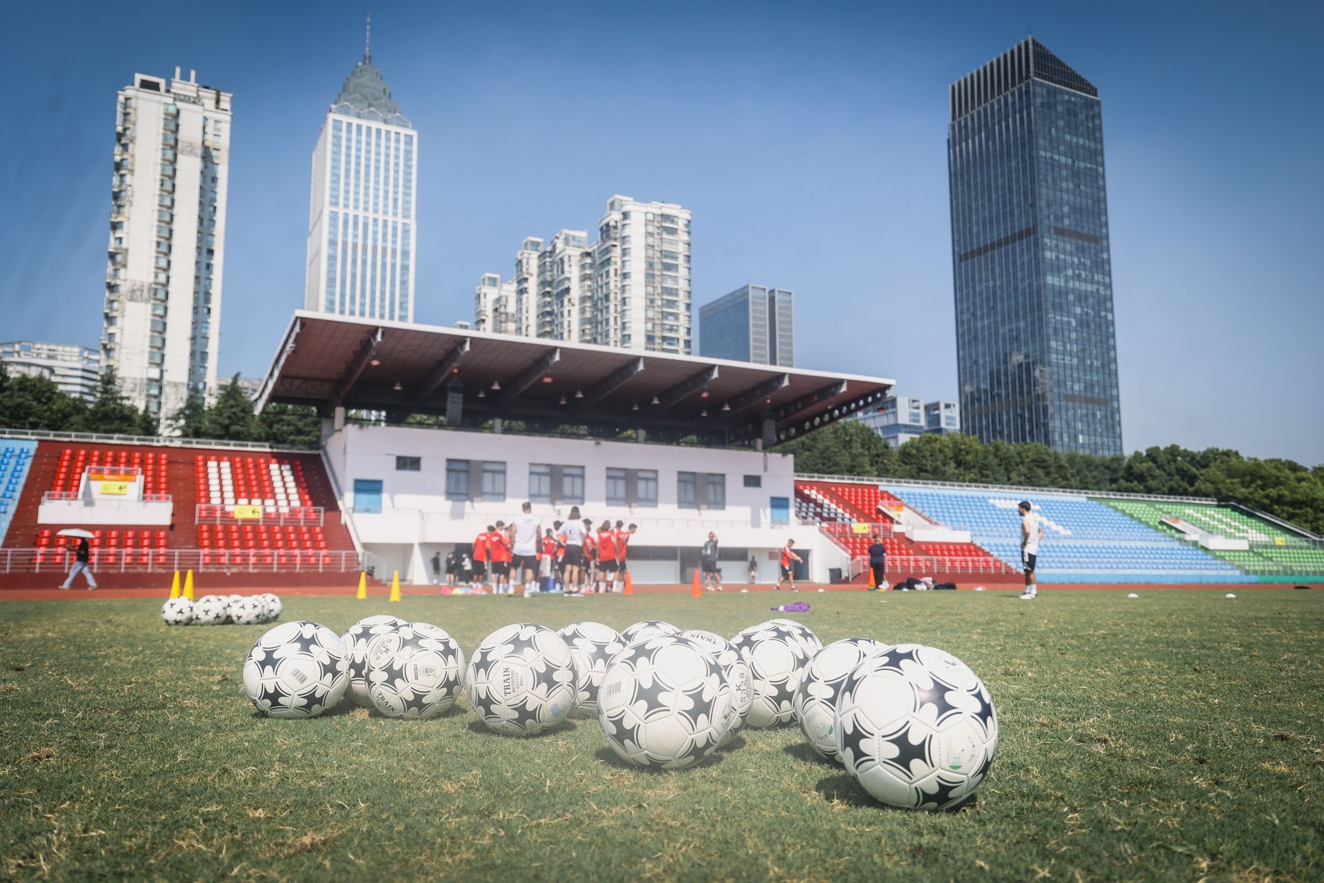 Ein Fußballfeld mit mehreren Bällen im Vordergrund, während eine Gruppe von Spielern im Hintergrund trainiert. Hochhäuser sind sichtbar.