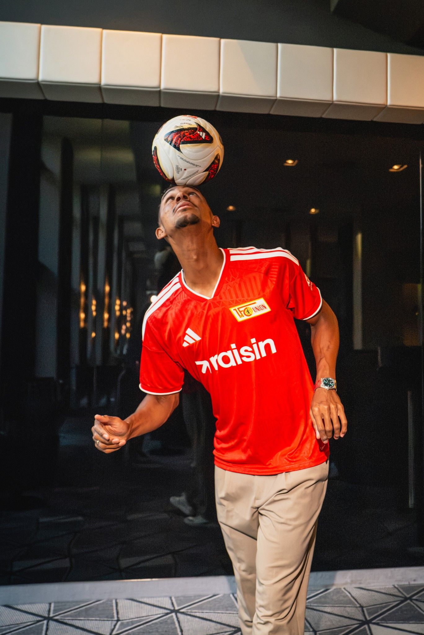 Ein Fußballspieler balanciert einen Ball auf dem Kopf, während er in die Kamera schaut und ein rotes Trikot trägt.