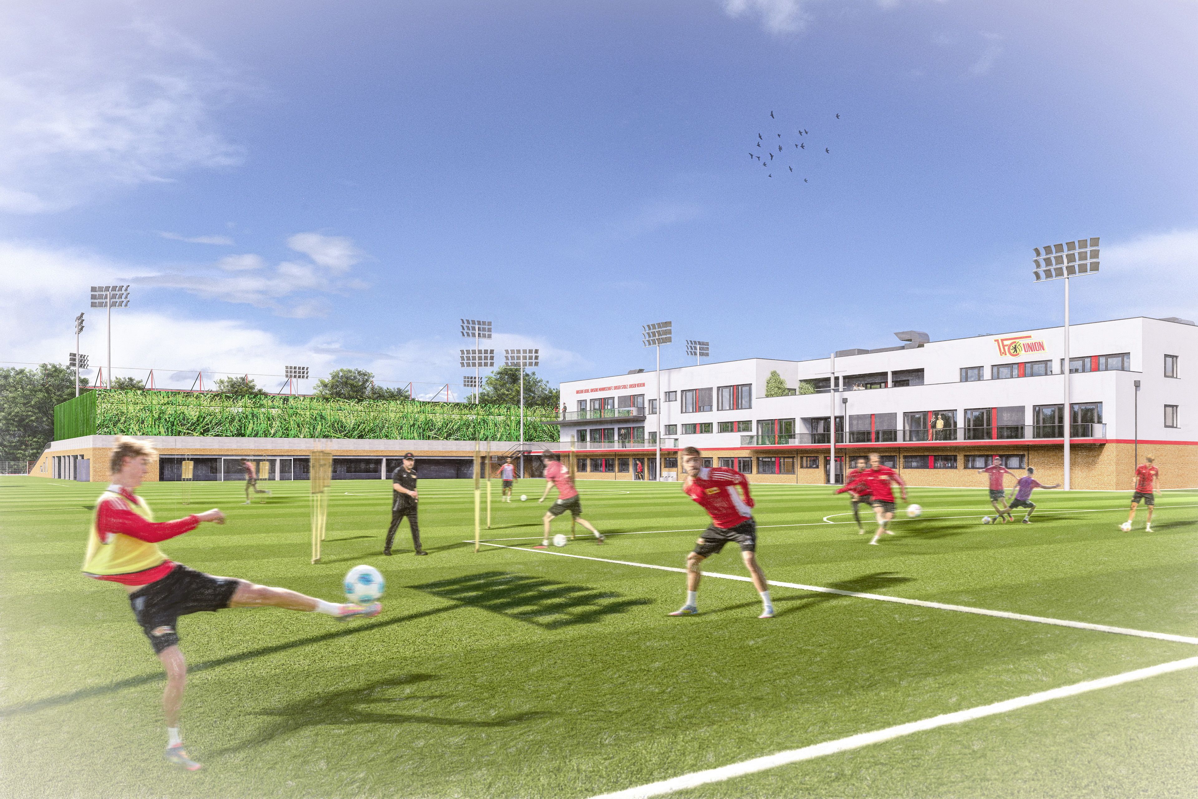 Fußballspieler trainieren auf einem grünen Spielfeld mit Toren und modernen Gebäuden im Hintergrund.