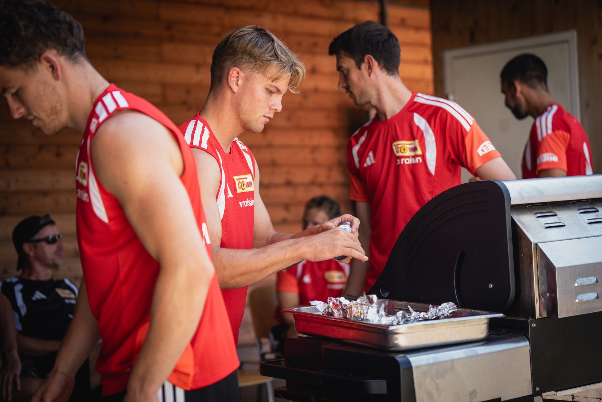 Drei männliche Sportler in roten Trikots bereiten Essen auf einem Grill vor, während ein weiterer Sportler im Hintergrund sitzt.