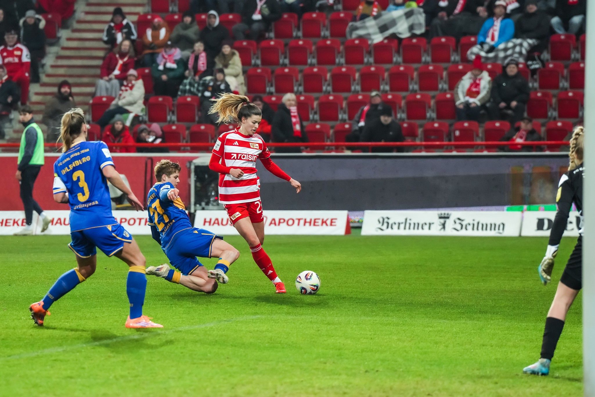 Eine Fußballspielerin in einem roten Trikot dribbelt den Ball, während eine Gegnerin im blauen Trikot sie zu Fall bringt.