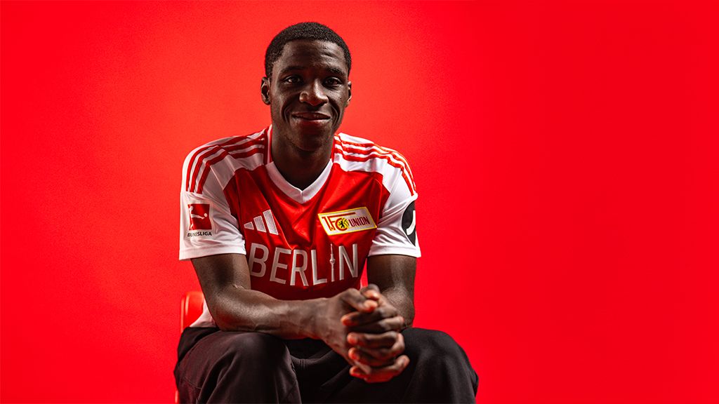 Ein junger Mann sitzt vor einem roten Hintergrund und trägt ein Fußballtrikot mit dem Logo von Union Berlin.