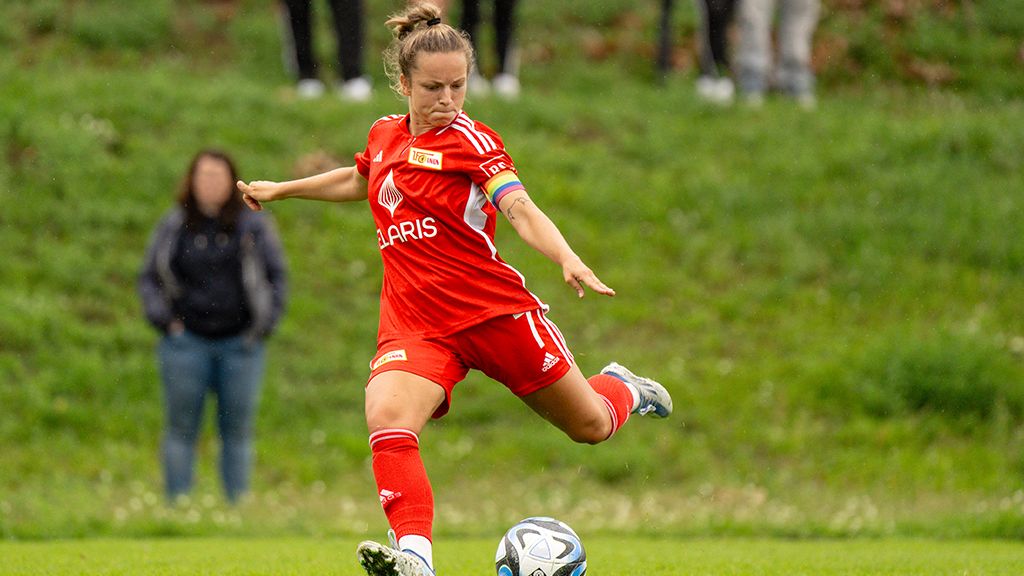 Eine Fußballspielerin in rotem Trikot tritt einen Ball auf einem Spielfeld. Zuschauer sind im Hintergrund sichtbar.