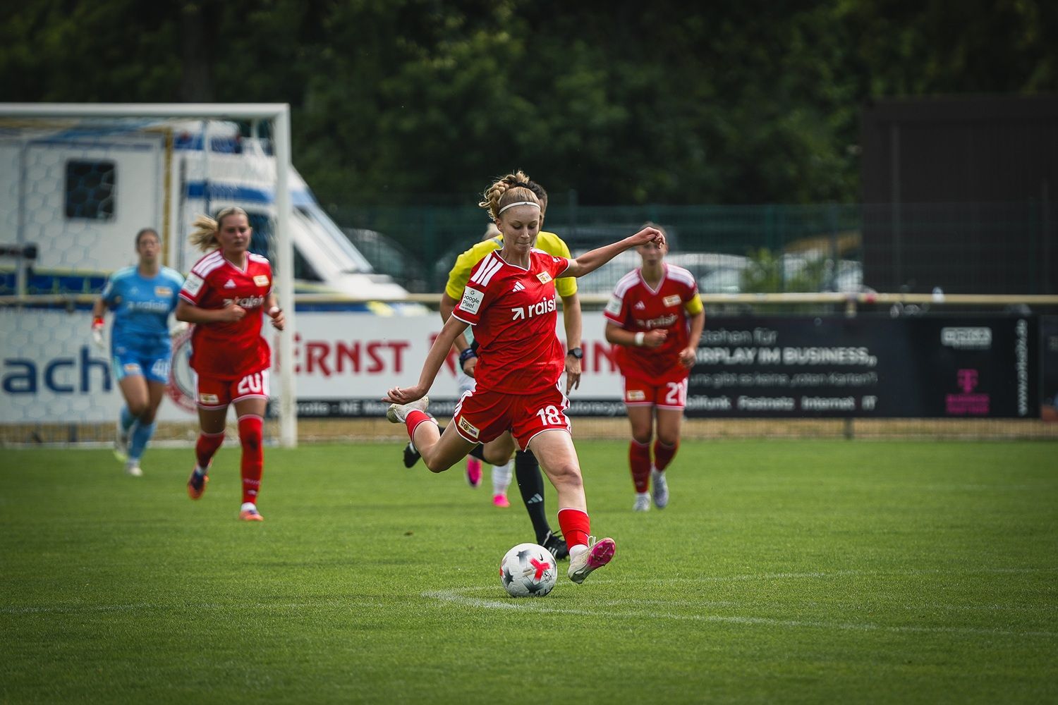Eine Fußballspielerin in roter Ausrüstung schießt den Ball, während Mitspielerinnen im Hintergrund stehen.