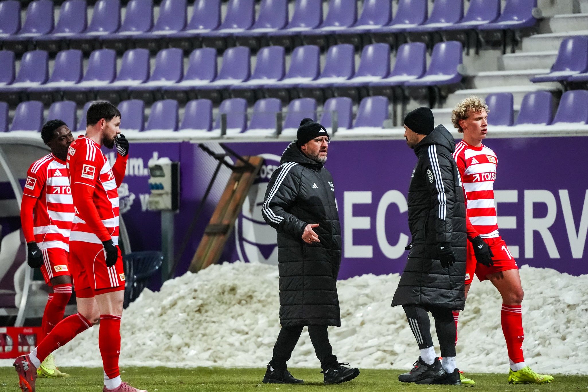 Fünf Fußballspieler in roten Trikots diskutieren mit einem Trainer in einem Stadion, während Schnee auf dem Spielfeld liegt.