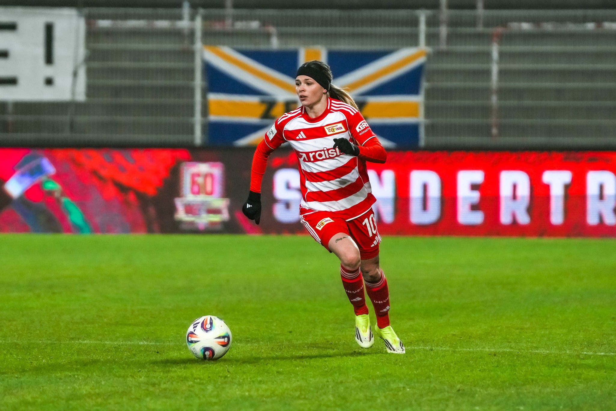 Eine Fußballspielerin in rot-weißem Trikot läuft über das Spielfeld, im Hintergrund ist eine Fahne und digitale Werbung sichtbar.