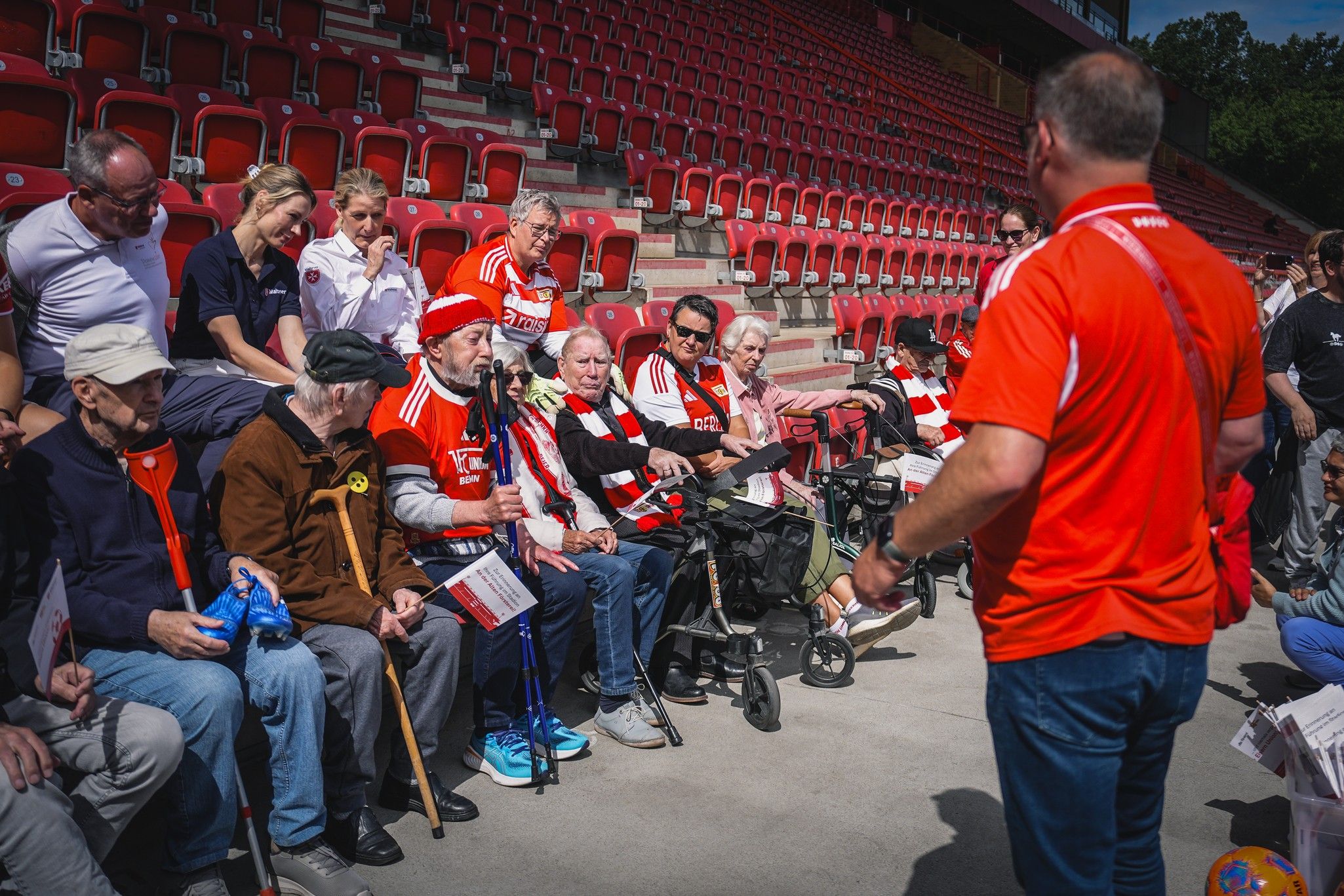 Ältere Fans in Stadion sitzend, hören einem redenden Mann in rotem Shirt zu, während sie Tickets in der Hand halten.