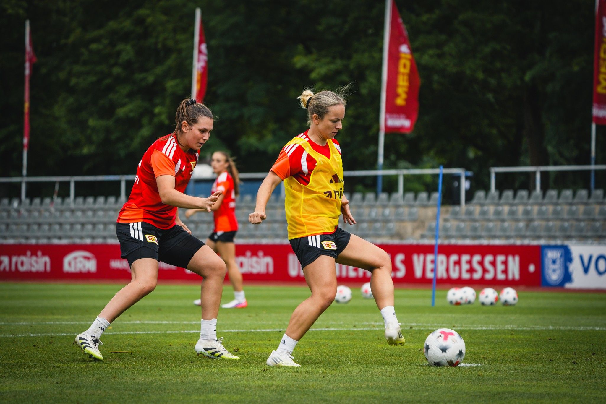 Zwei Fußballspielerinnen trainieren auf einem Rasenplatz, eine trägt ein gelbes Trikot, die andere ein rotes.