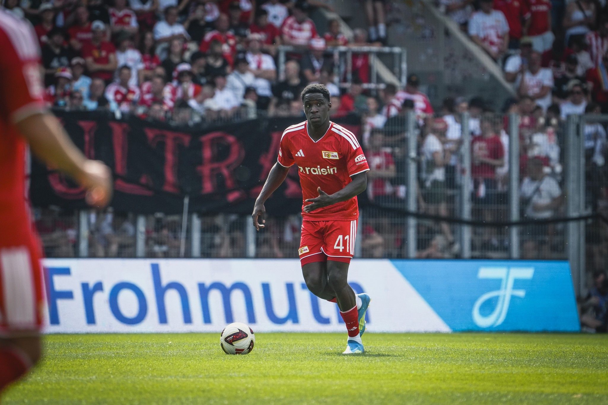Ein Fußballspieler in rotem Trikot dribbelt den Ball auf dem Spielfeld, hinter ihm Fans in einer jubelnden Kulisse.