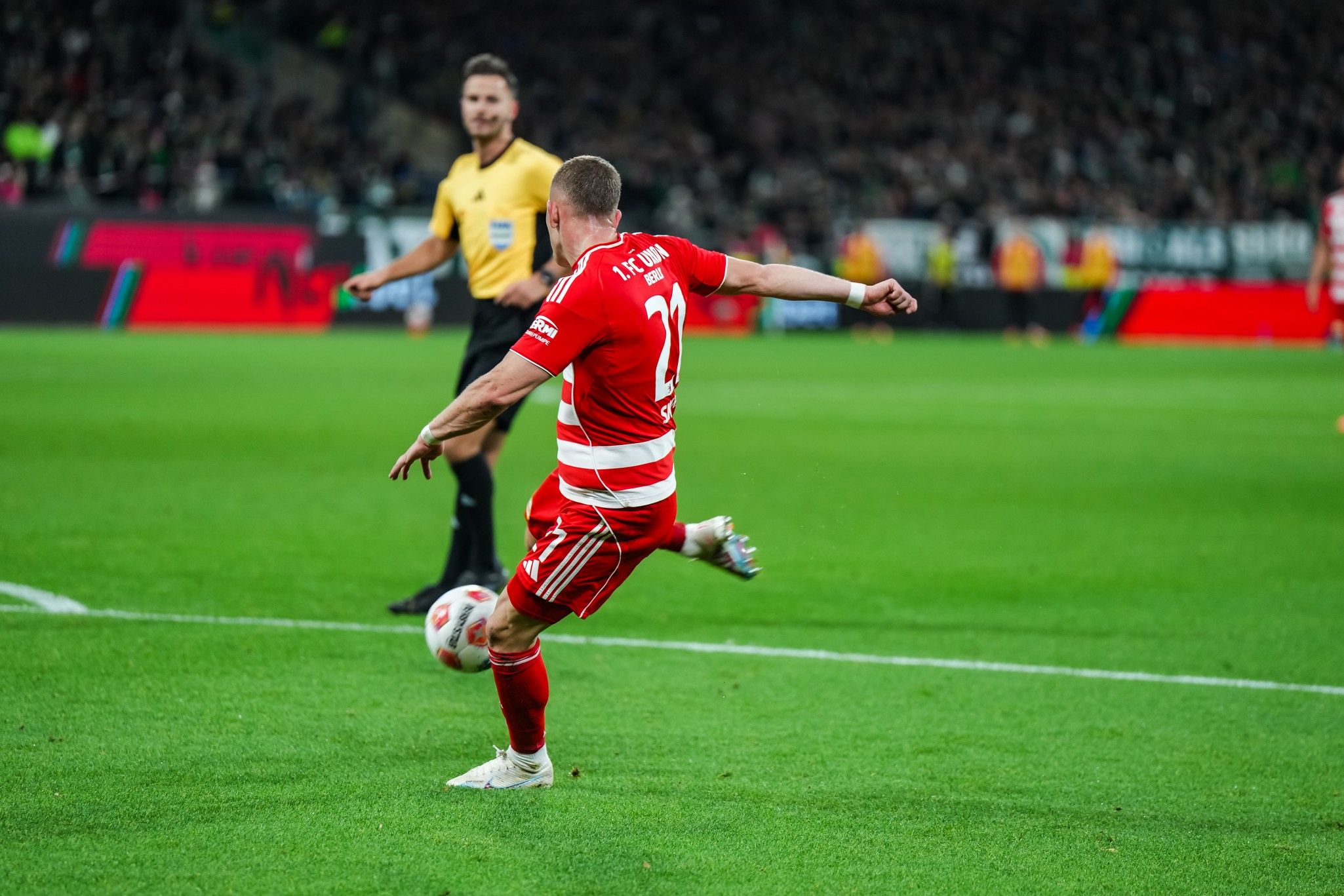 Spieler in rotem Trikot führt einen Eckball aus, während der Schiedsrichter im Hintergrund zuschaut.